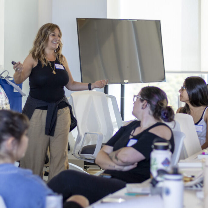 A woman wearing brown pants and a black top presents to a group of adult learners at a CRLP event.